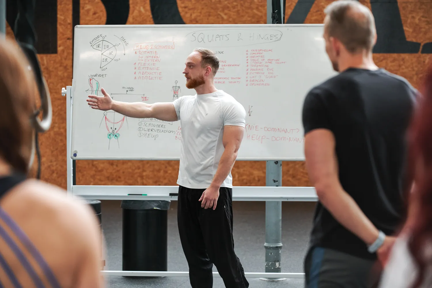 Docent legt op het whiteboard het schema voor squats en hinges uit aan de groep tijdens een praktijkdag.