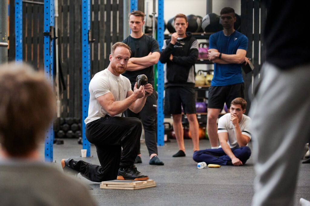 Docent demonstreert de techniek van een oefening aan cursisten tijdens een live praktijkdag in de sportschool.